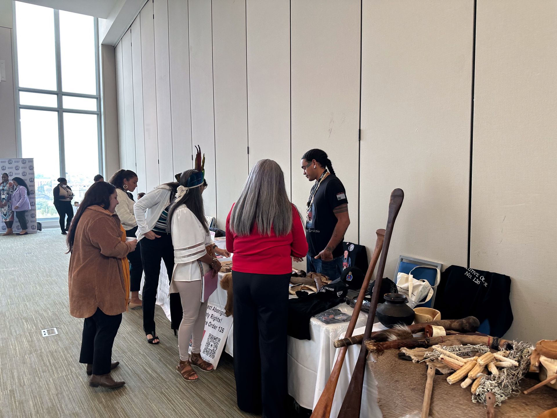 People browse a vendor's table at an event. Displayed items include crafts and tools. A bright, interior setting.