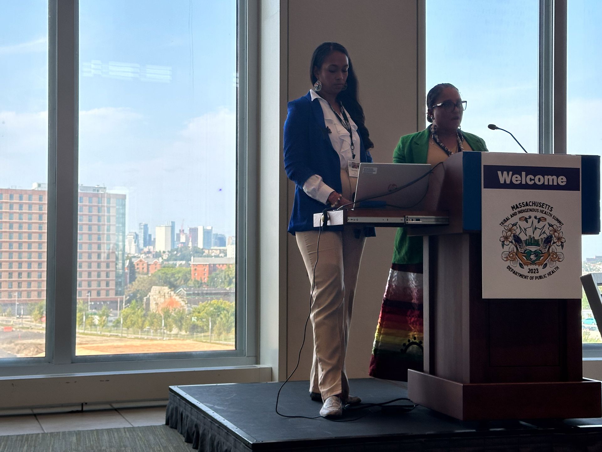 Two women at a podium, one presenting with a laptop. City view background.