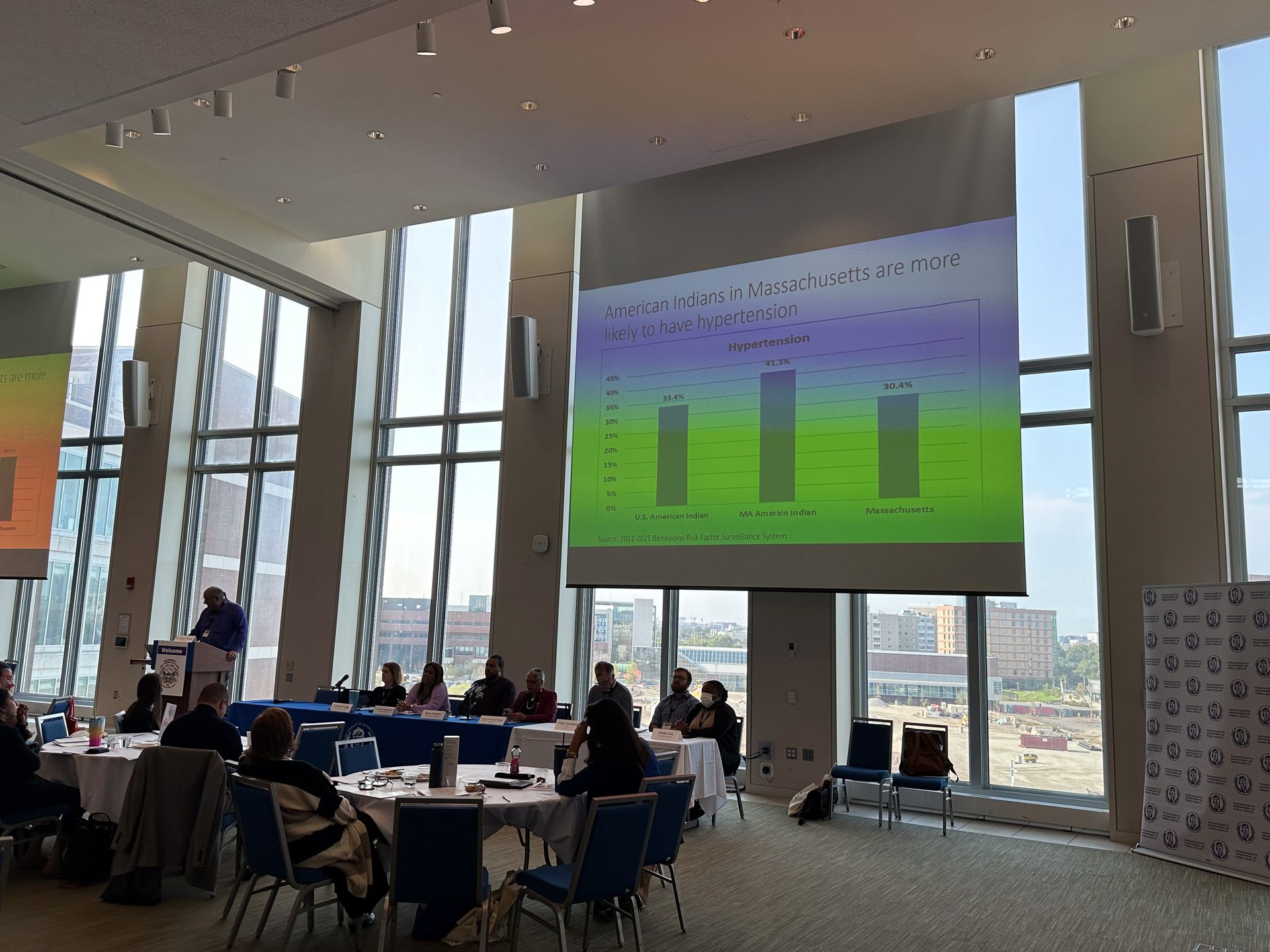 A conference presentation: speaker at podium, audience seated at tables, projected bar graph, large windows.
