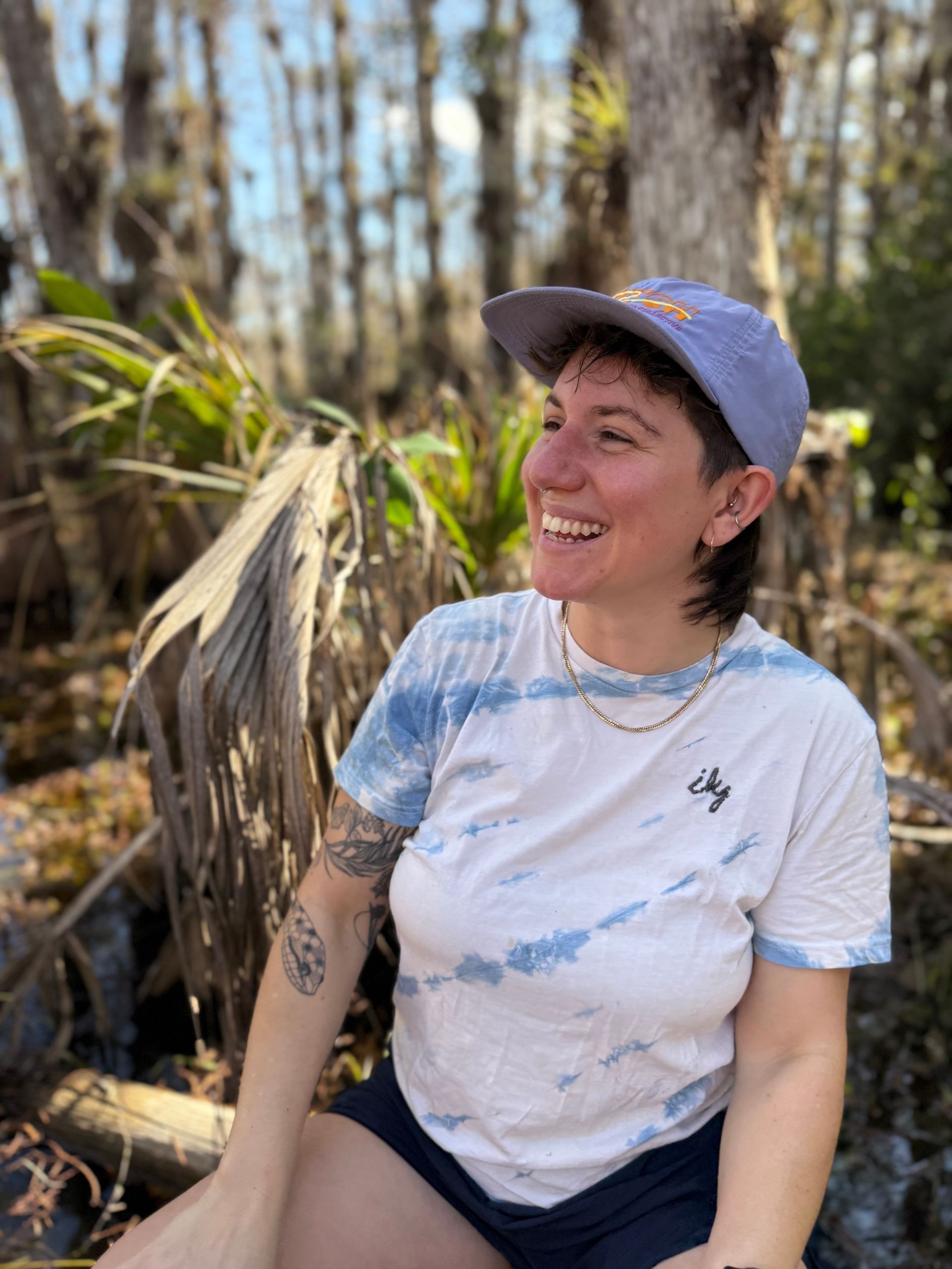 Person smiling, wearing a blue tie-dye shirt and hat, outdoors in a swamp-like setting.