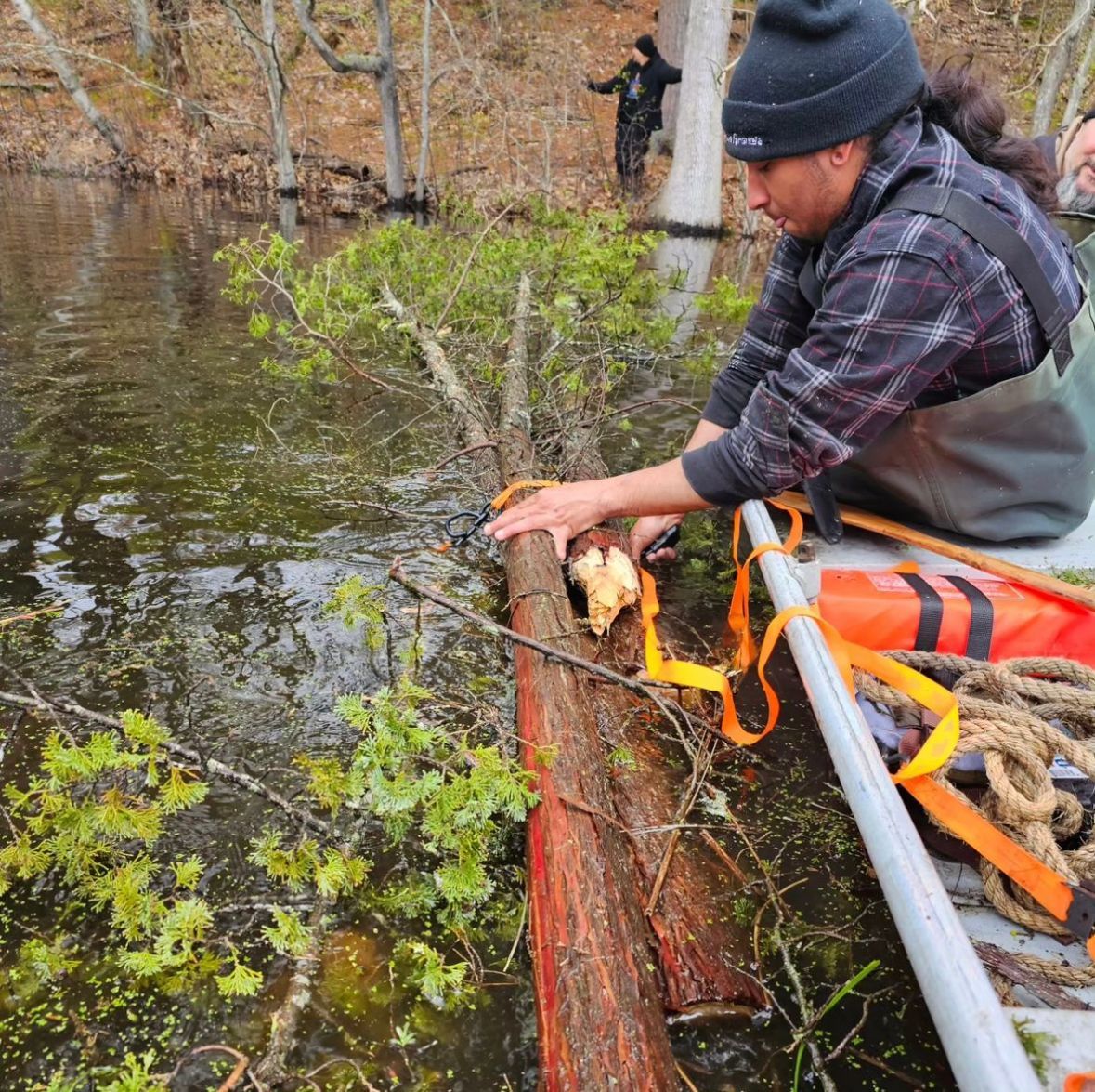Person in a boat secures a log in a body of water with another person in the background.