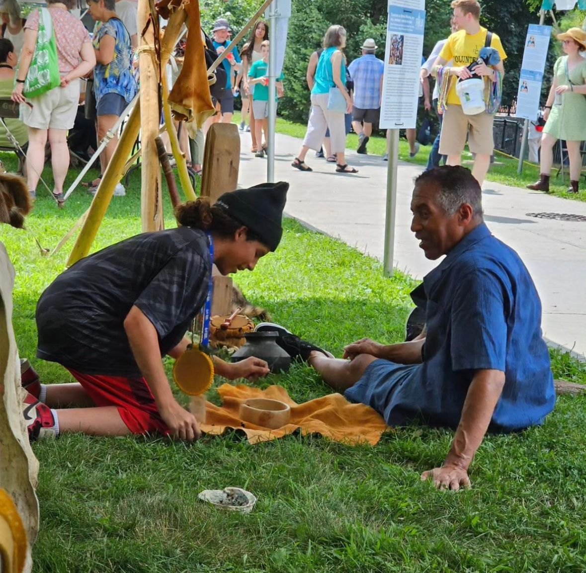 Two people sit on grass, one tending to the other's foot. Outdoor setting with onlookers.