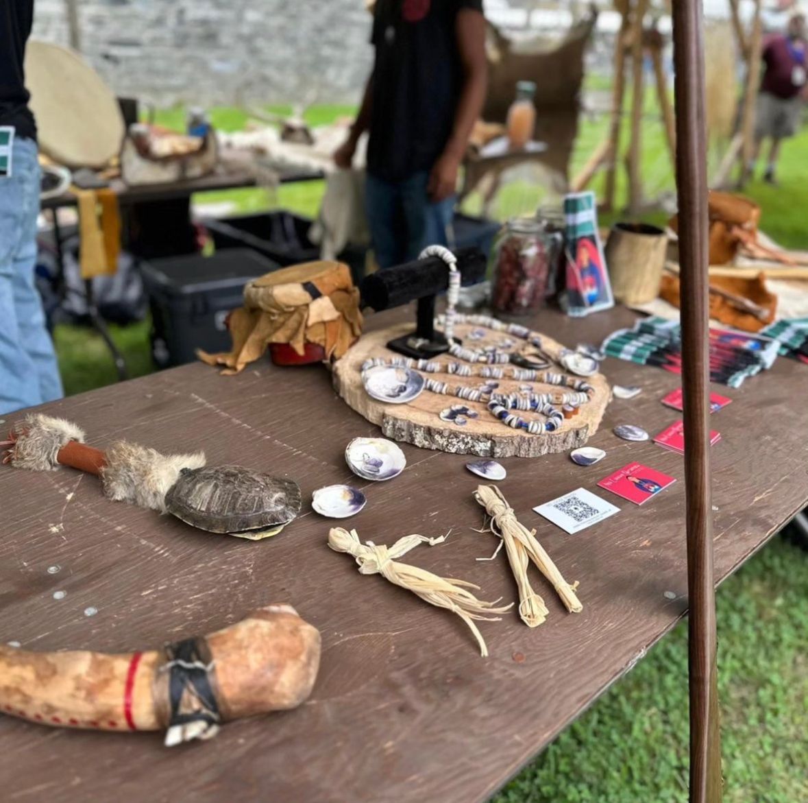 Table of crafts: turtle shell, jewelry, herbs, with people and drums in the background.
