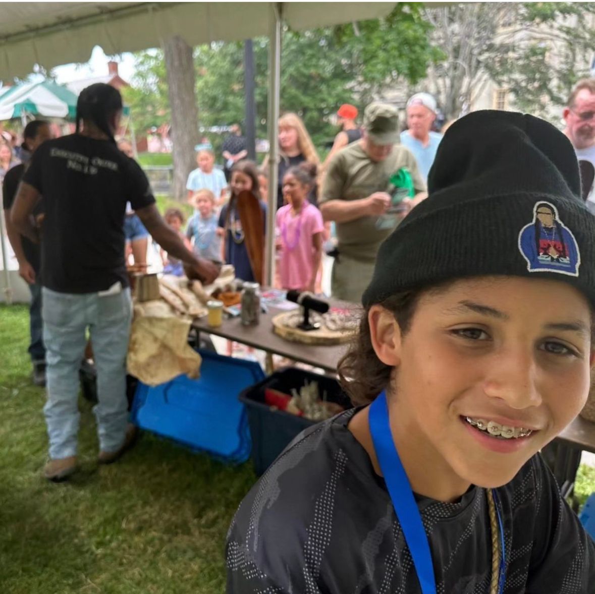 Boy in beanie smiles at the camera, wearing braces and a lanyard, at an outdoor event.