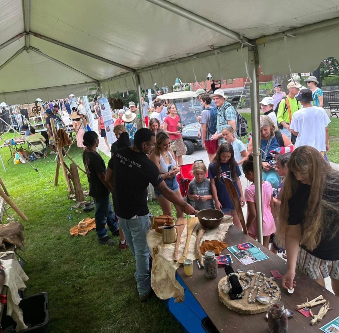 People at outdoor display under tent; man showing objects to children, other people nearby.