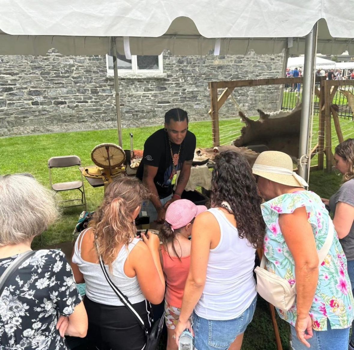 People at a craft fair booth under a tent. A person is demonstrating, and others are watching.