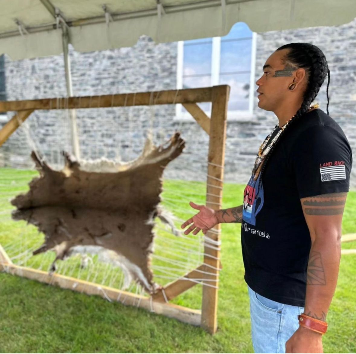 Man points at a stretched animal hide in front of a stone building.