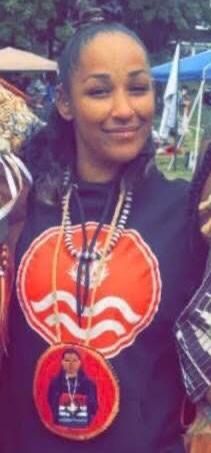 Woman wearing a black top with an orange and white circular design and a beaded necklace, smiling in an outdoor setting.