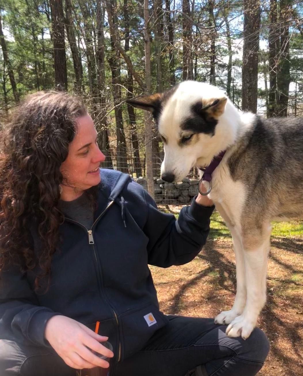 Woman seated, petting a dog with black and white markings; outdoors, sunny day.