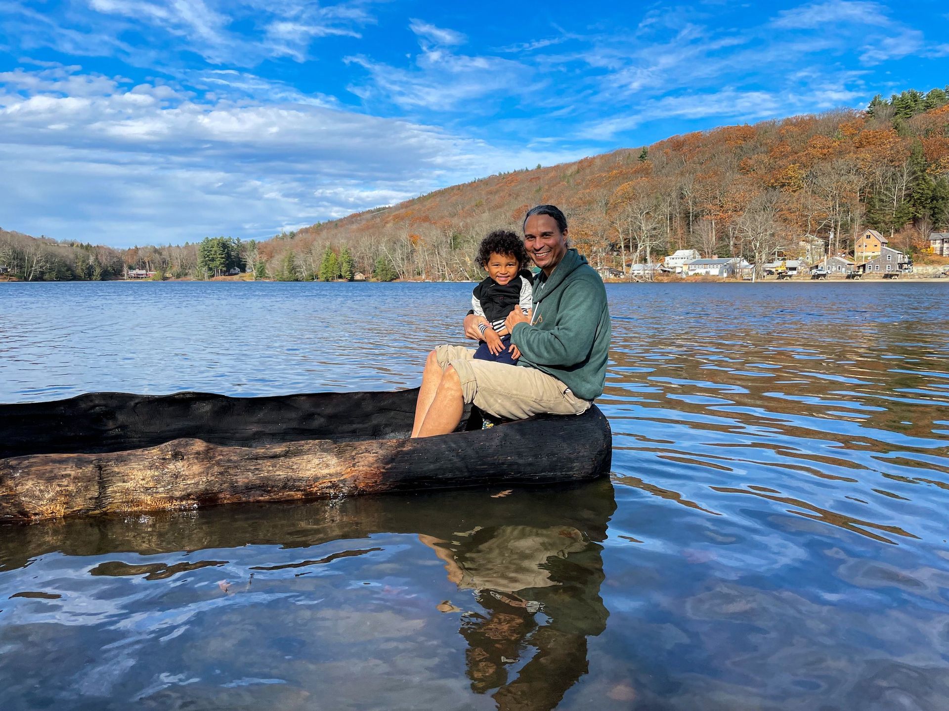 Man and child sit in a makeshift floating log on a lake; fall foliage in the background.
