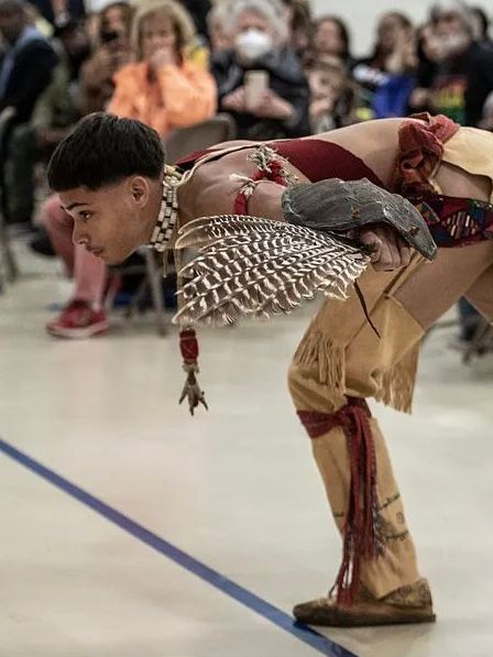 A dancer in Native American regalia, bent over, performing on a stage. Audience in the background.
