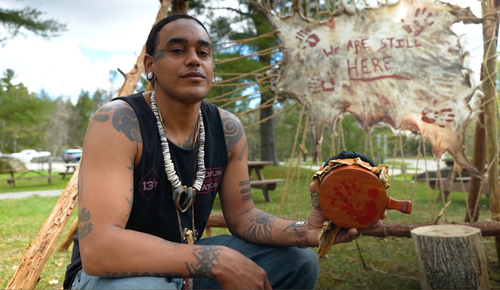 Man with tattoos, holding a drum, sits near a sign that says 