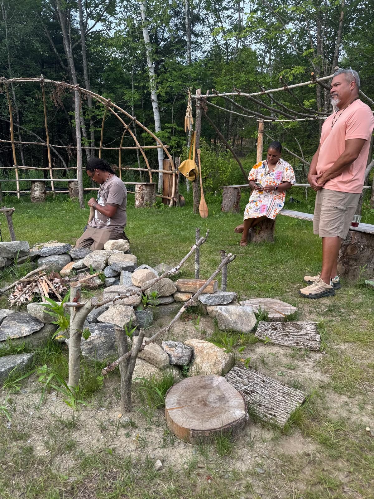 People near a rock fire pit and wooden structure in a grassy outdoor setting.