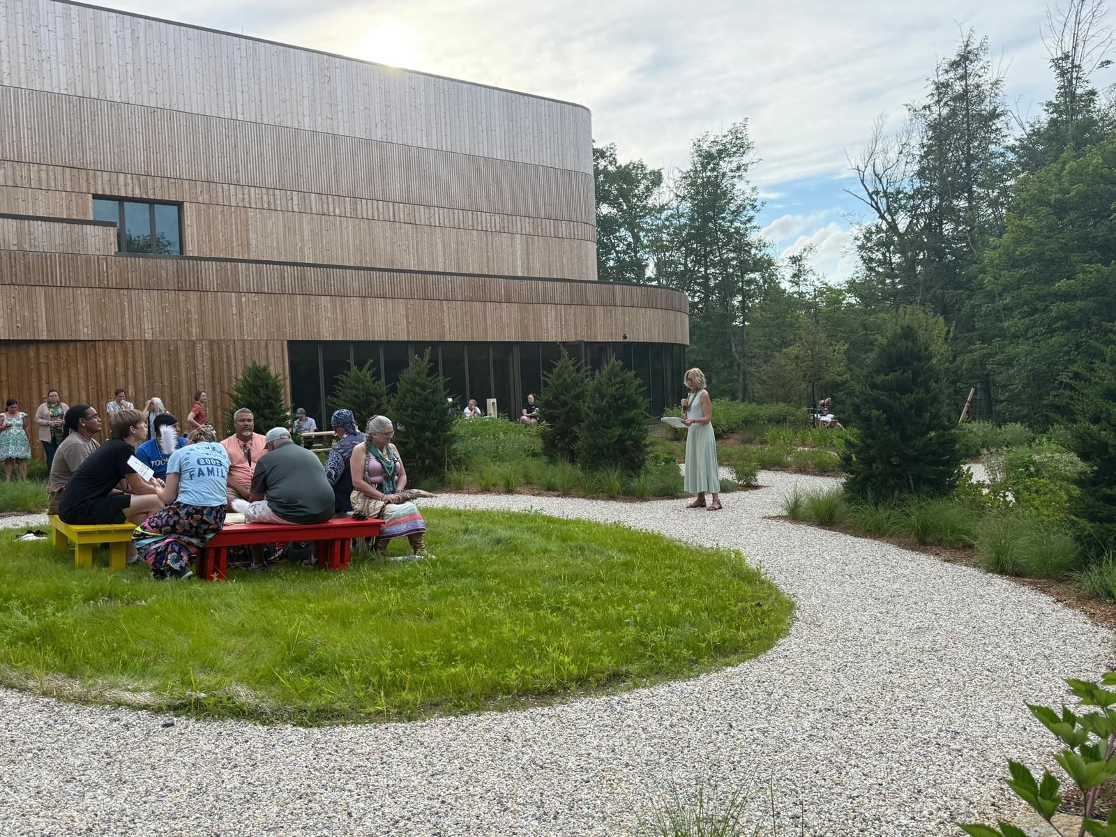 Group seated outdoors, listening to a woman speaking near a modern wooden building.