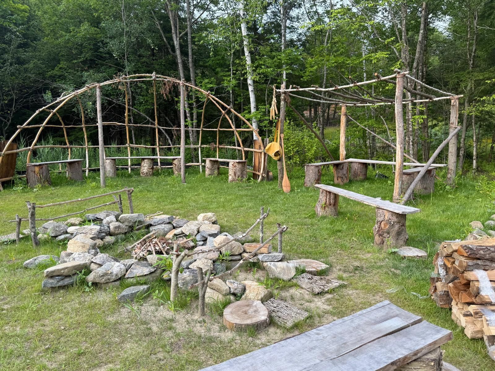 Campground with wooden structures, benches, fire pit, and firewood. Green grass and trees in the background.