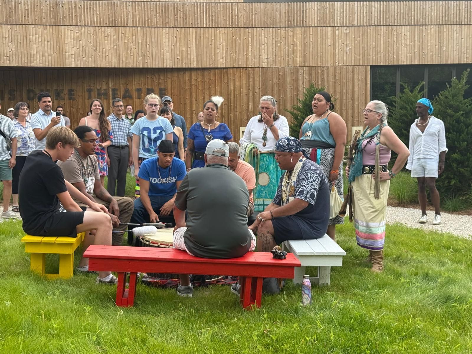 Group of people gathered outdoors, some singing near a building. Red benches and colorful leis are visible.