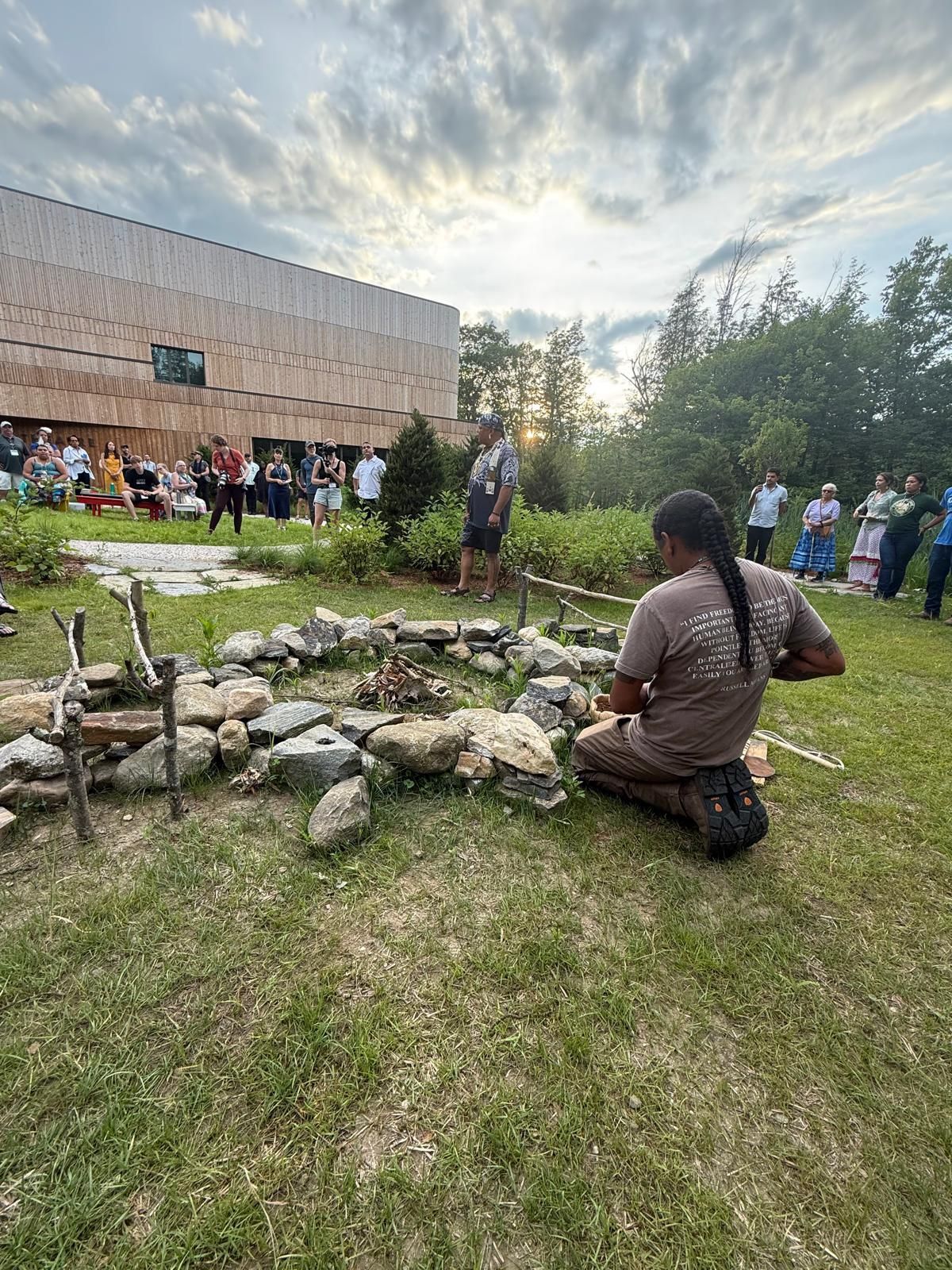 Person kneeling by a stone fire pit, lighting it. Others watch from the grass near a wooden building.