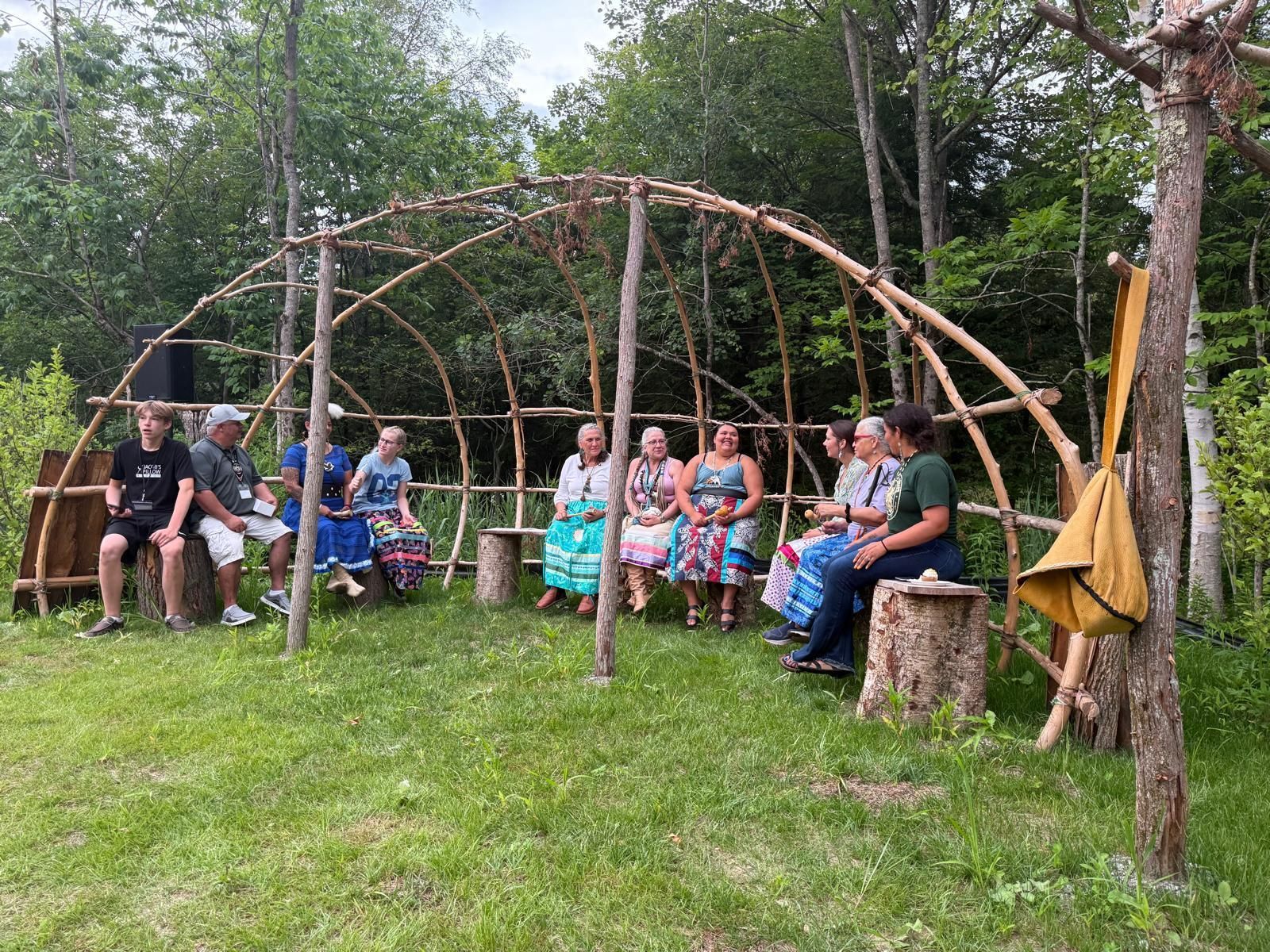 People seated under a wooden arbor in a grassy outdoor setting, possibly a gathering or event.