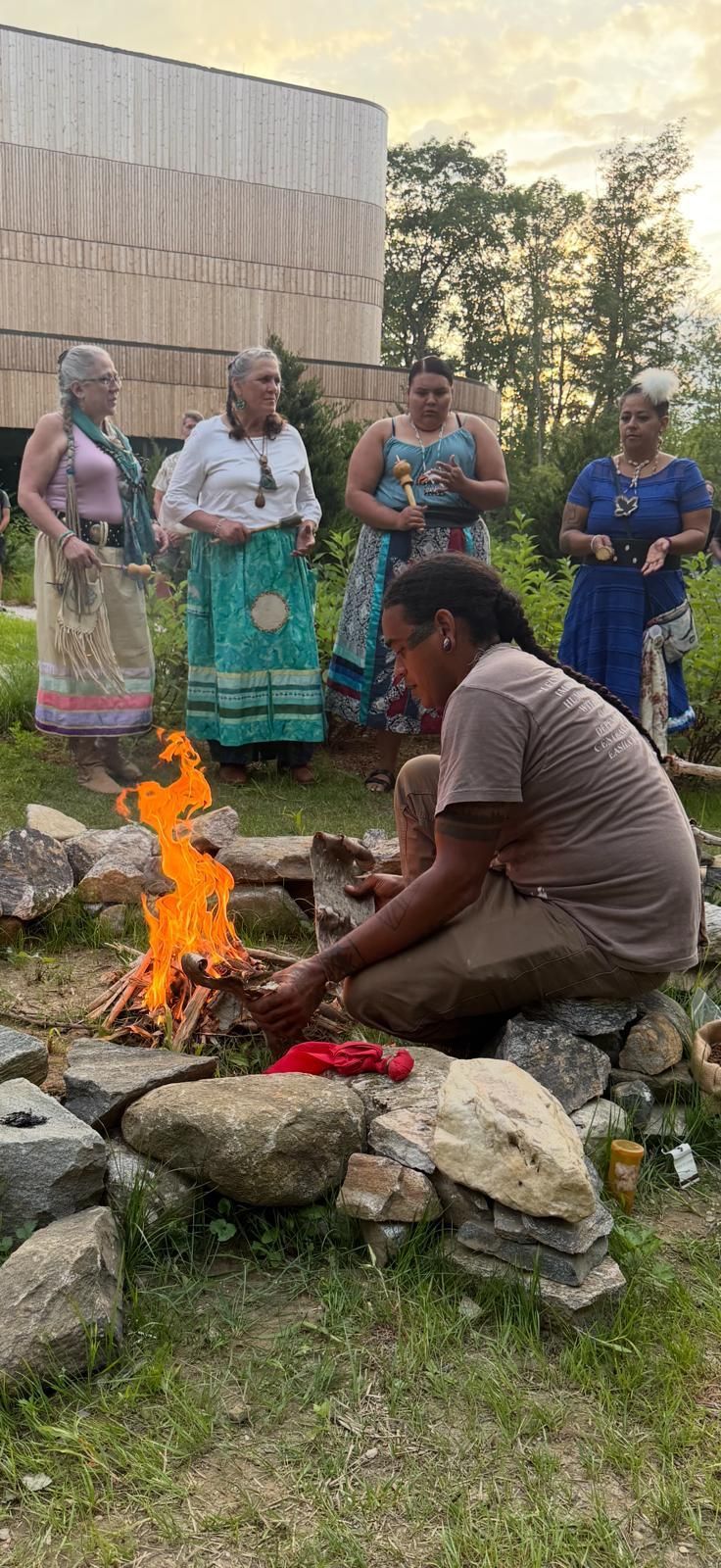 A person kneels near a fire. Four women stand behind. They are outside next to a building.