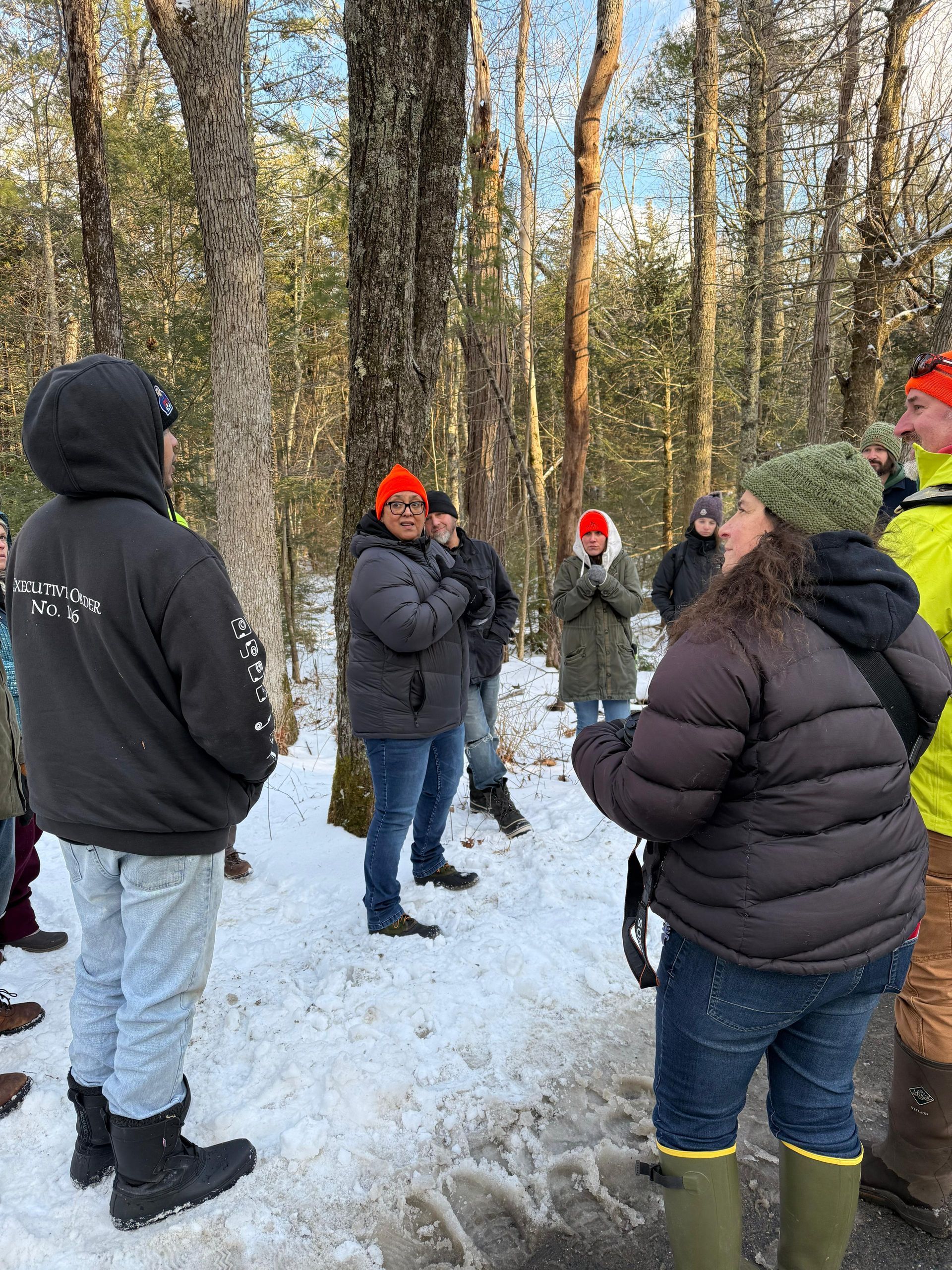 A group of people in winter clothing stand in a snowy forest, some wearing orange hats.