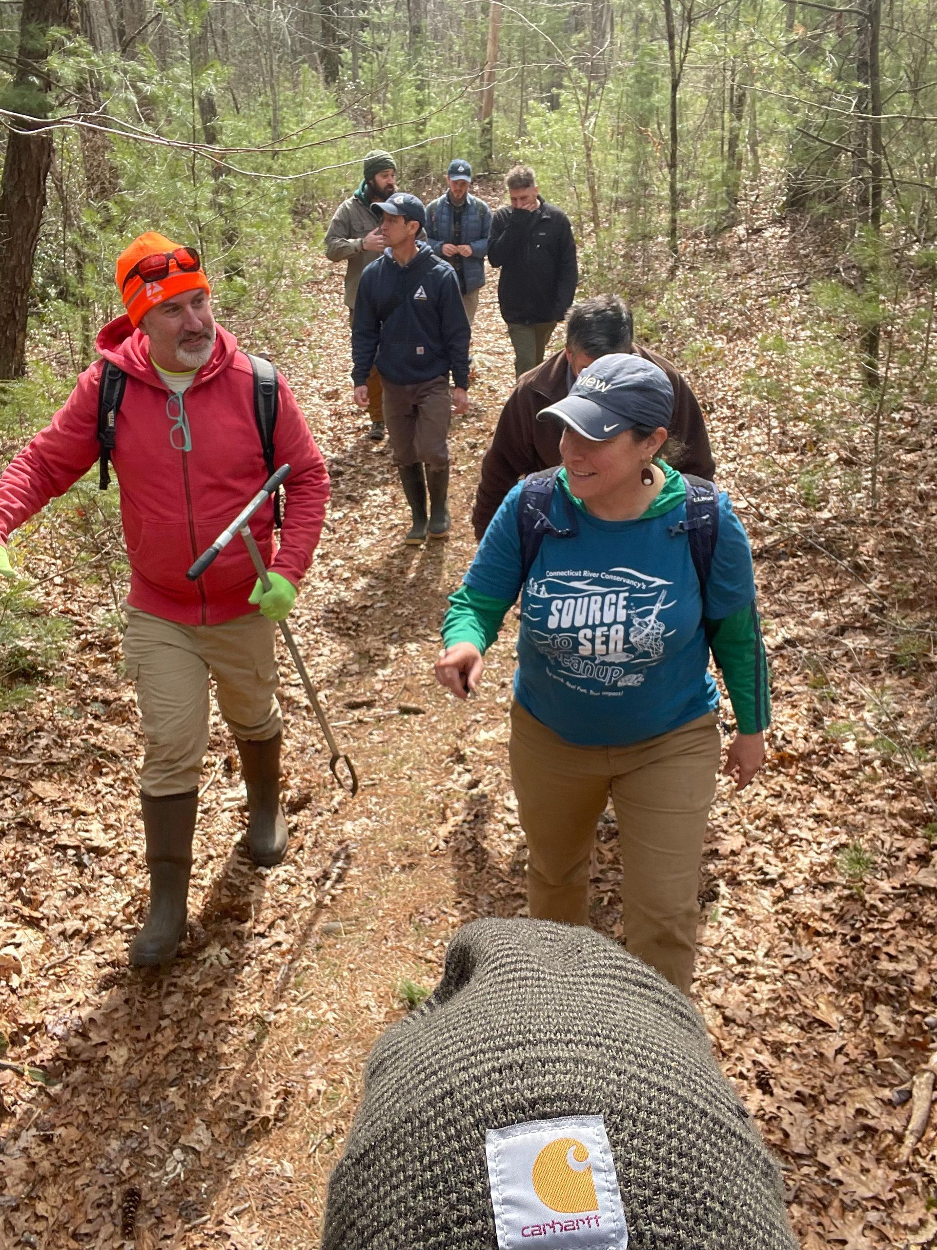 People hiking on a leaf-covered forest trail. Person in front wears work gloves, others in jackets and hats.