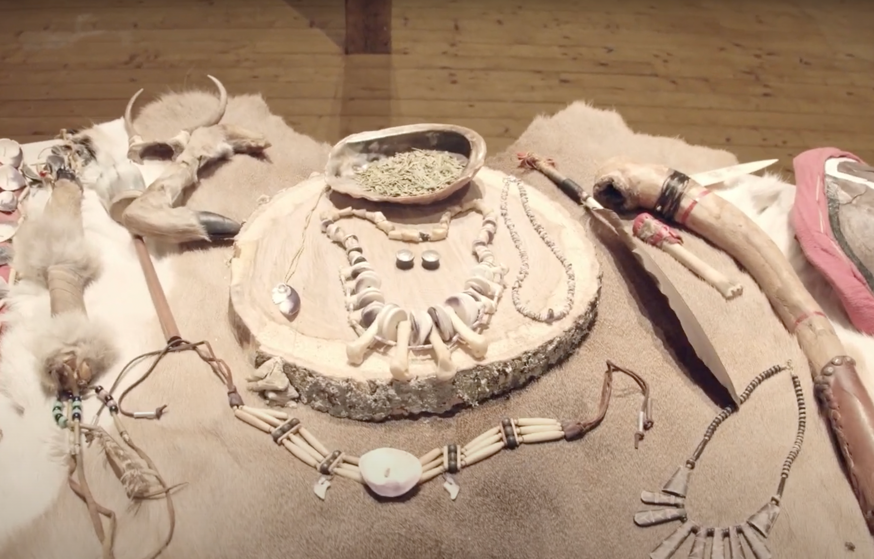 Artifacts displayed on fur: carved wood, bone jewelry, skulls, and a bowl with green items.