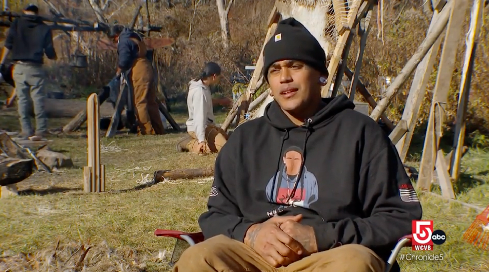 Man in black hoodie outdoors, speaking. Wooden structures and people in background.