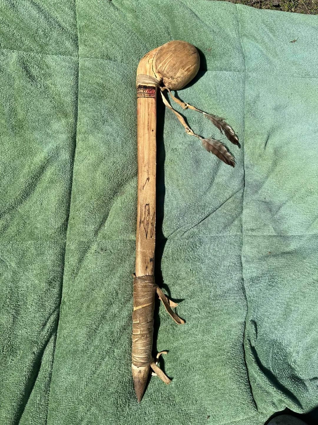Native American war club with a rounded head, wooden shaft, and attached feathers, resting on green fabric.