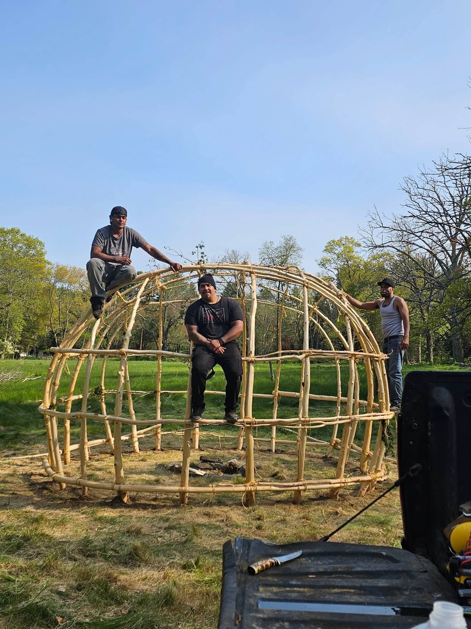 Three people building a wooden dome structure outdoors on a sunny day.