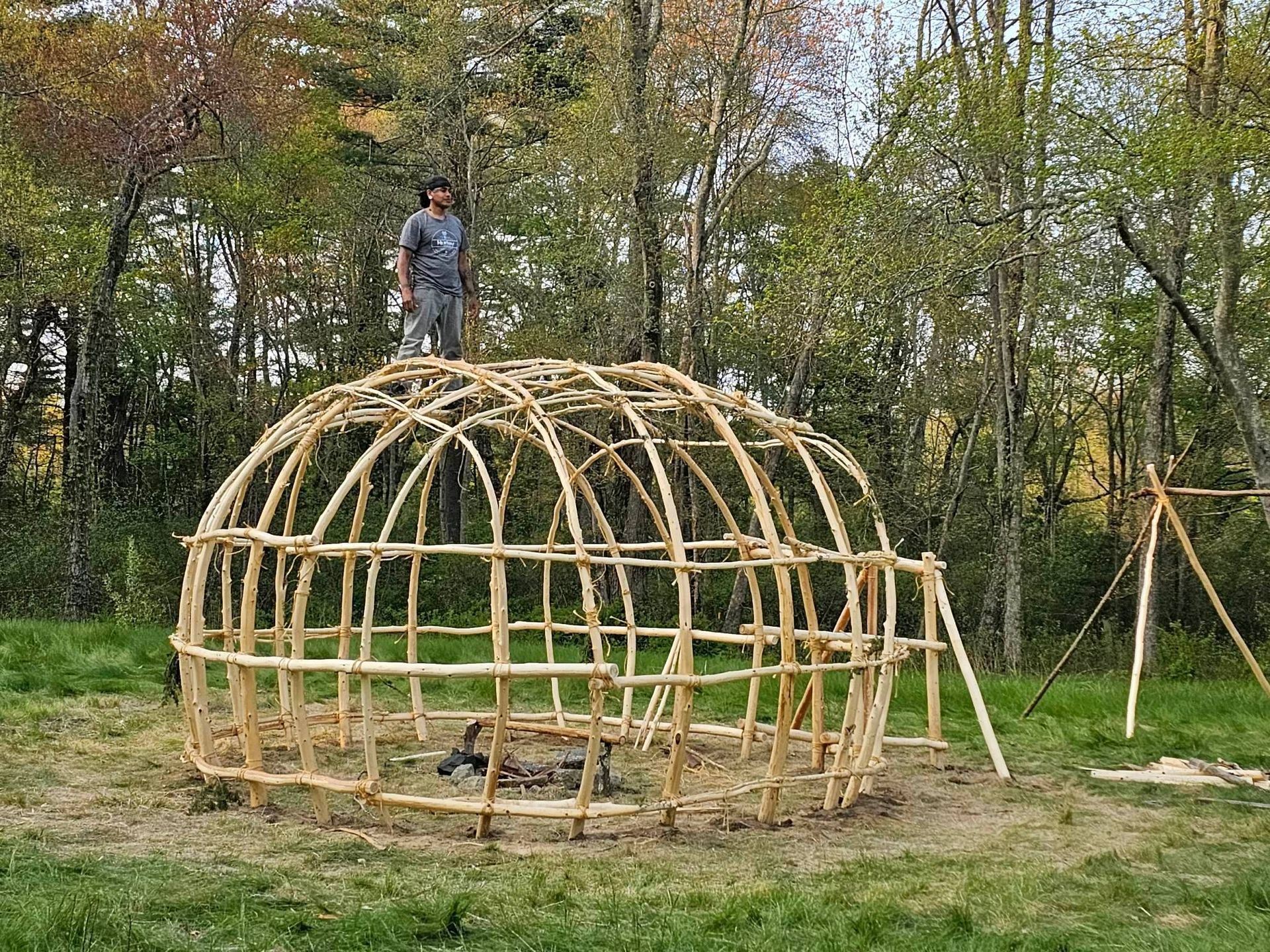 Person standing atop a wooden dome-shaped frame in a grassy field. Trees in the background.