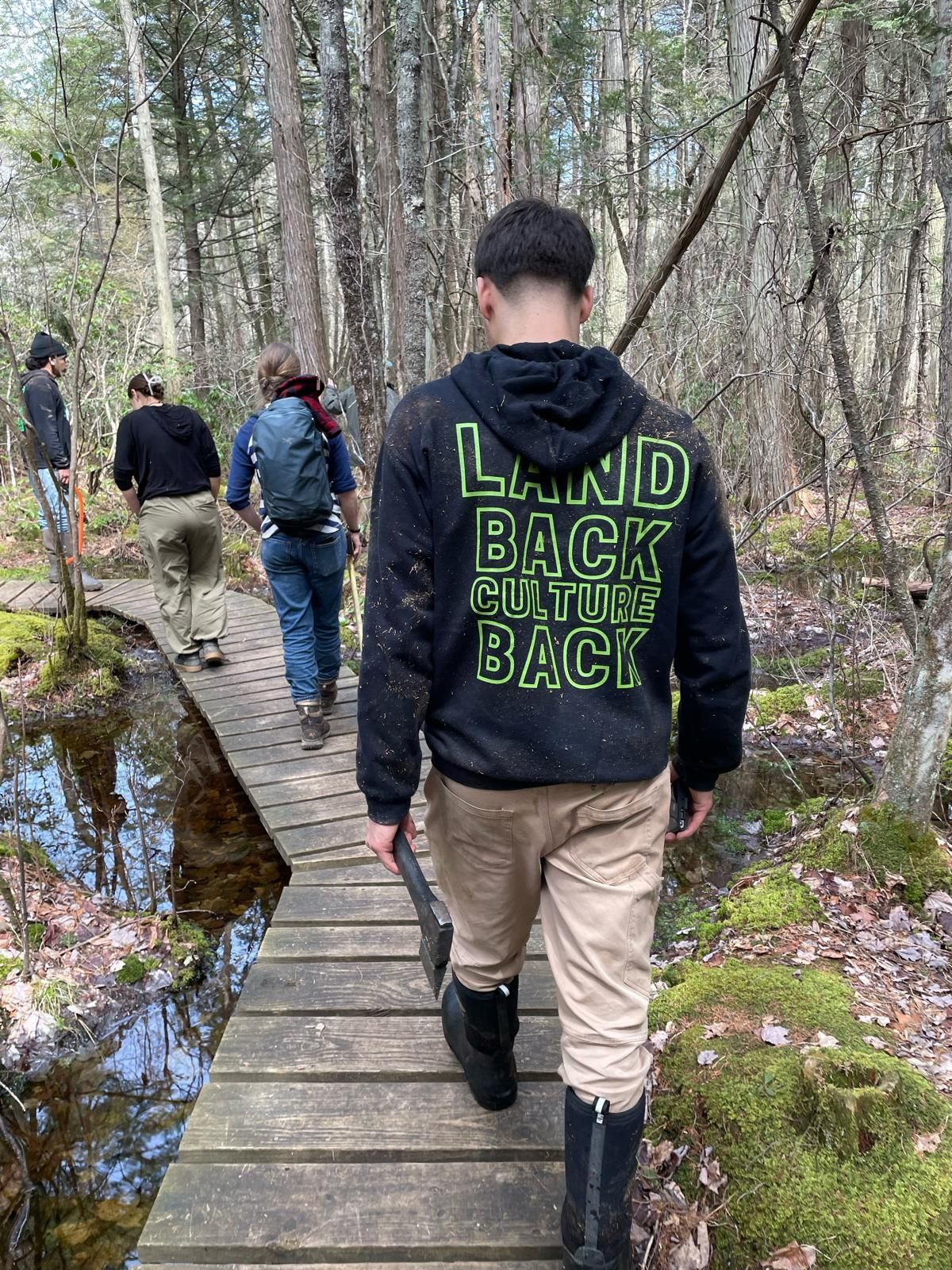 People walking on a wooden boardwalk through a wooded area. One wears a black hoodie with green text reading 