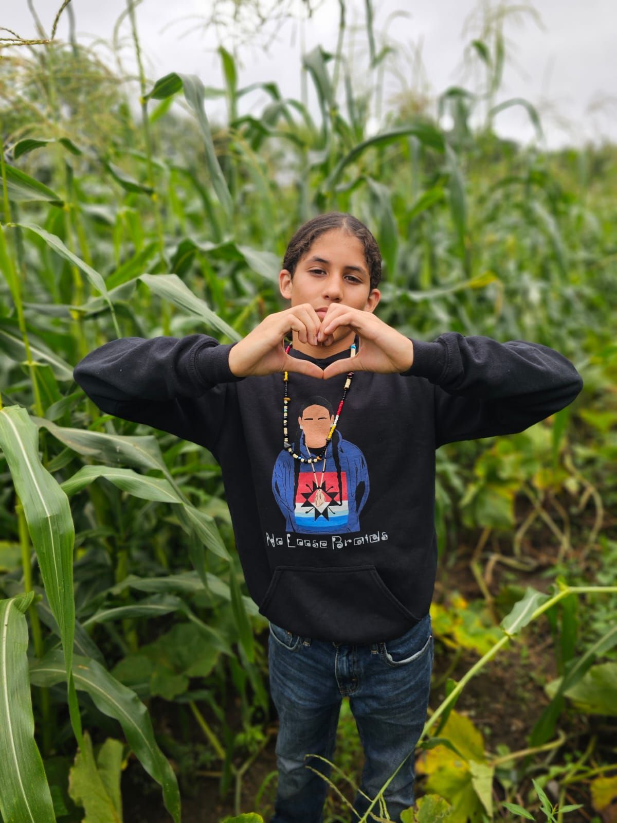 Person in black hoodie making a heart shape with their hands, standing in a cornfield.