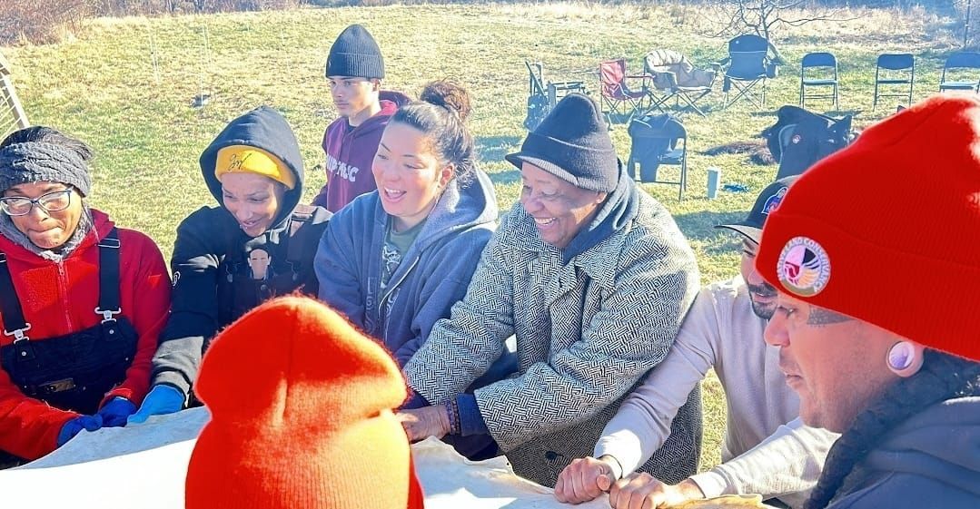 Group of people gathered around a table outdoors, some wearing hats and coats. They appear to be interacting and smiling.