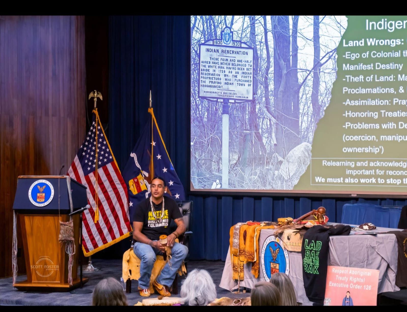 Man speaking at a podium with US and NY flags. Screen displays 