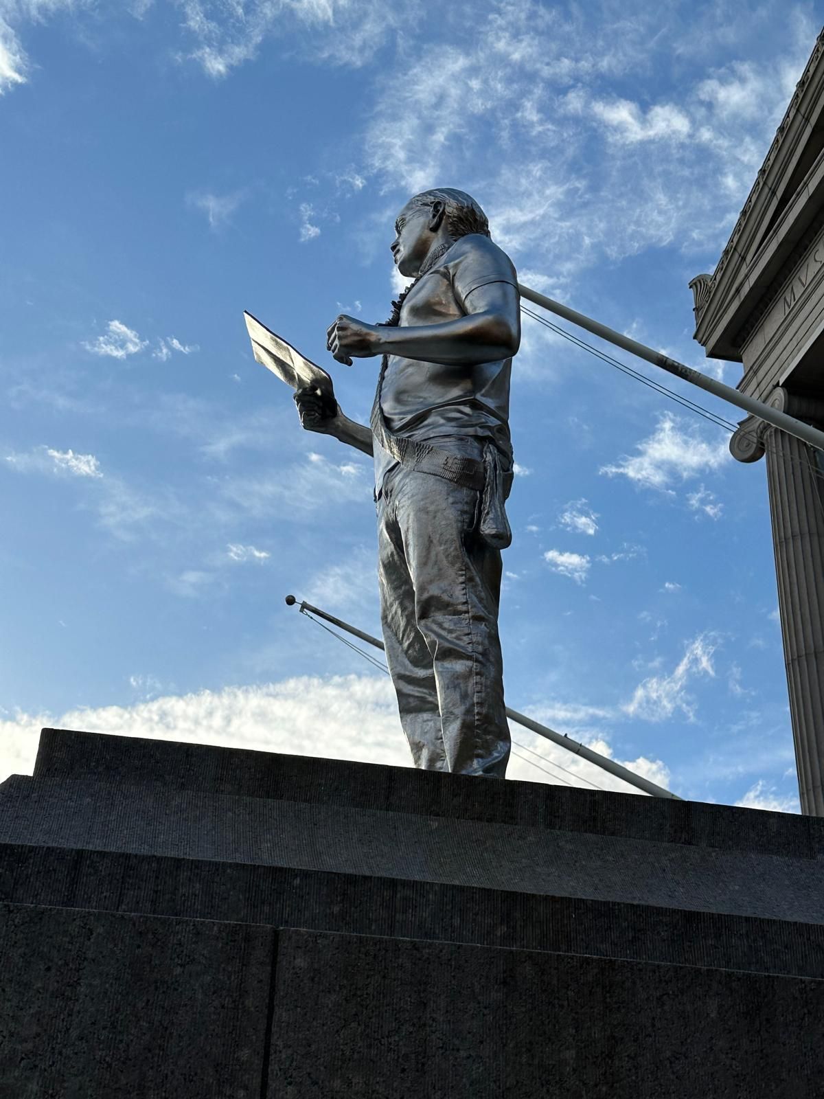 Silver statue of a person holding a tablet, standing atop steps, against a blue sky.