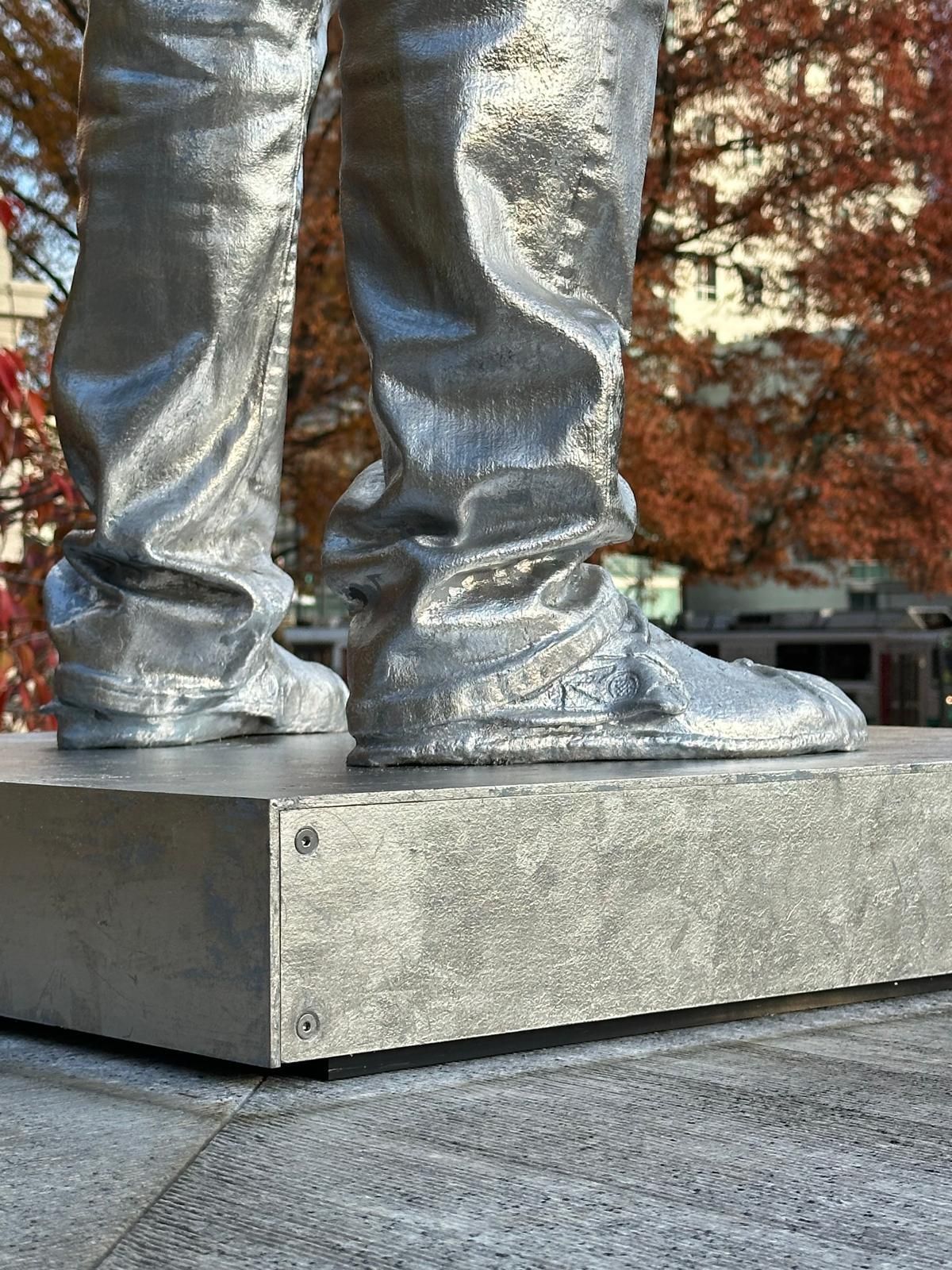 Silver statue's legs and feet on a rectangular base; autumn foliage in the background.