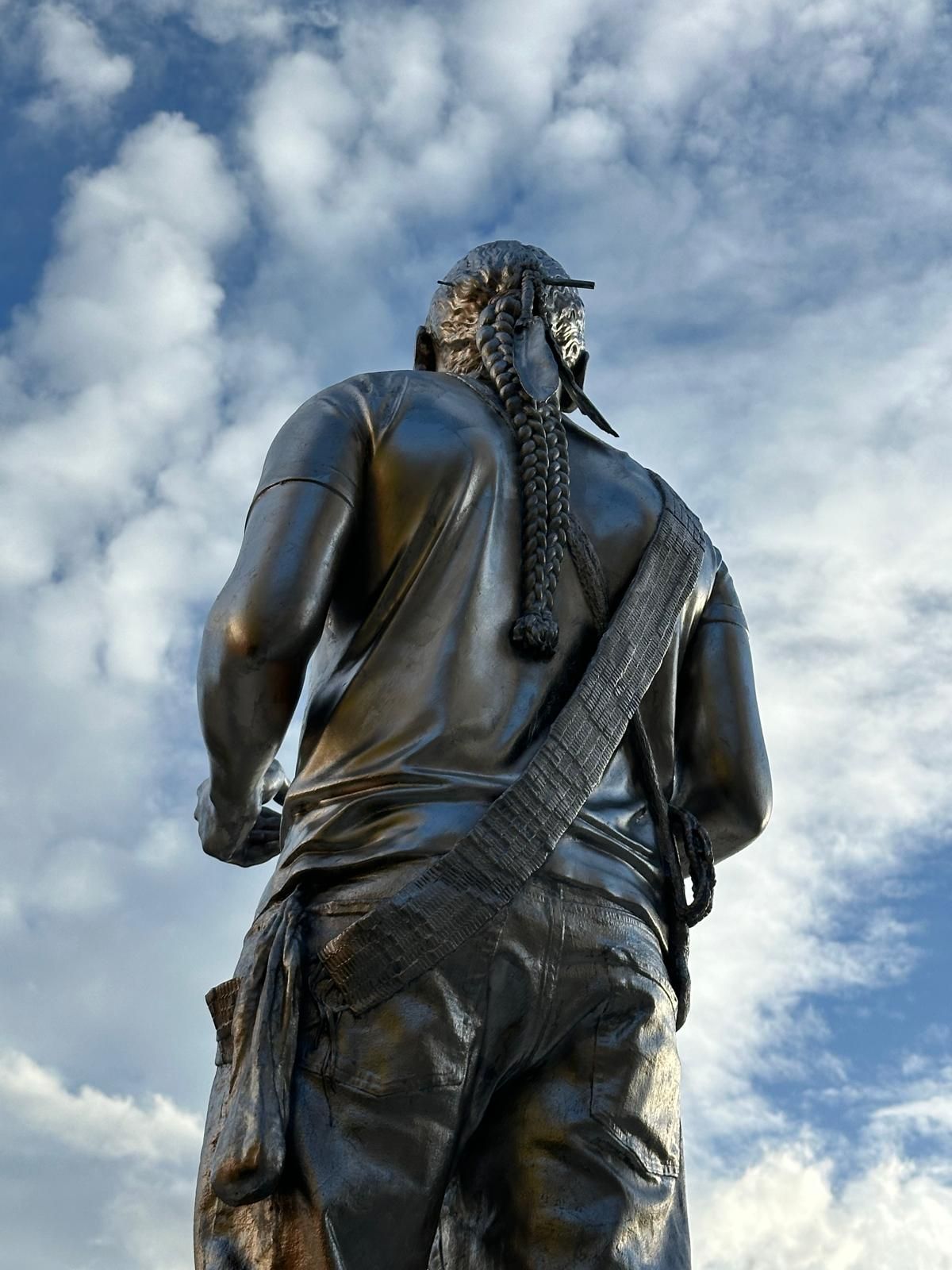 Bronze statue of a person, back view, wearing a long braid and strap across the body.
