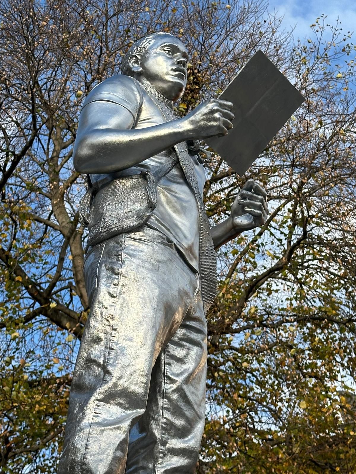 Silver statue of a person holding a tablet, standing outdoors near a tree with fall foliage.