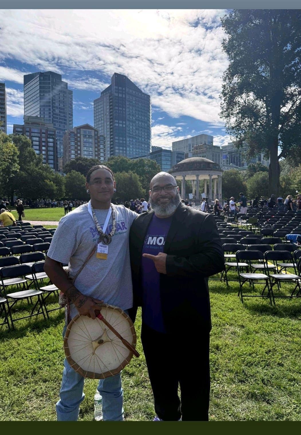 Two men pose outdoors; one holds a drum. Skyscrapers and seated crowd in background. Sunny day.
