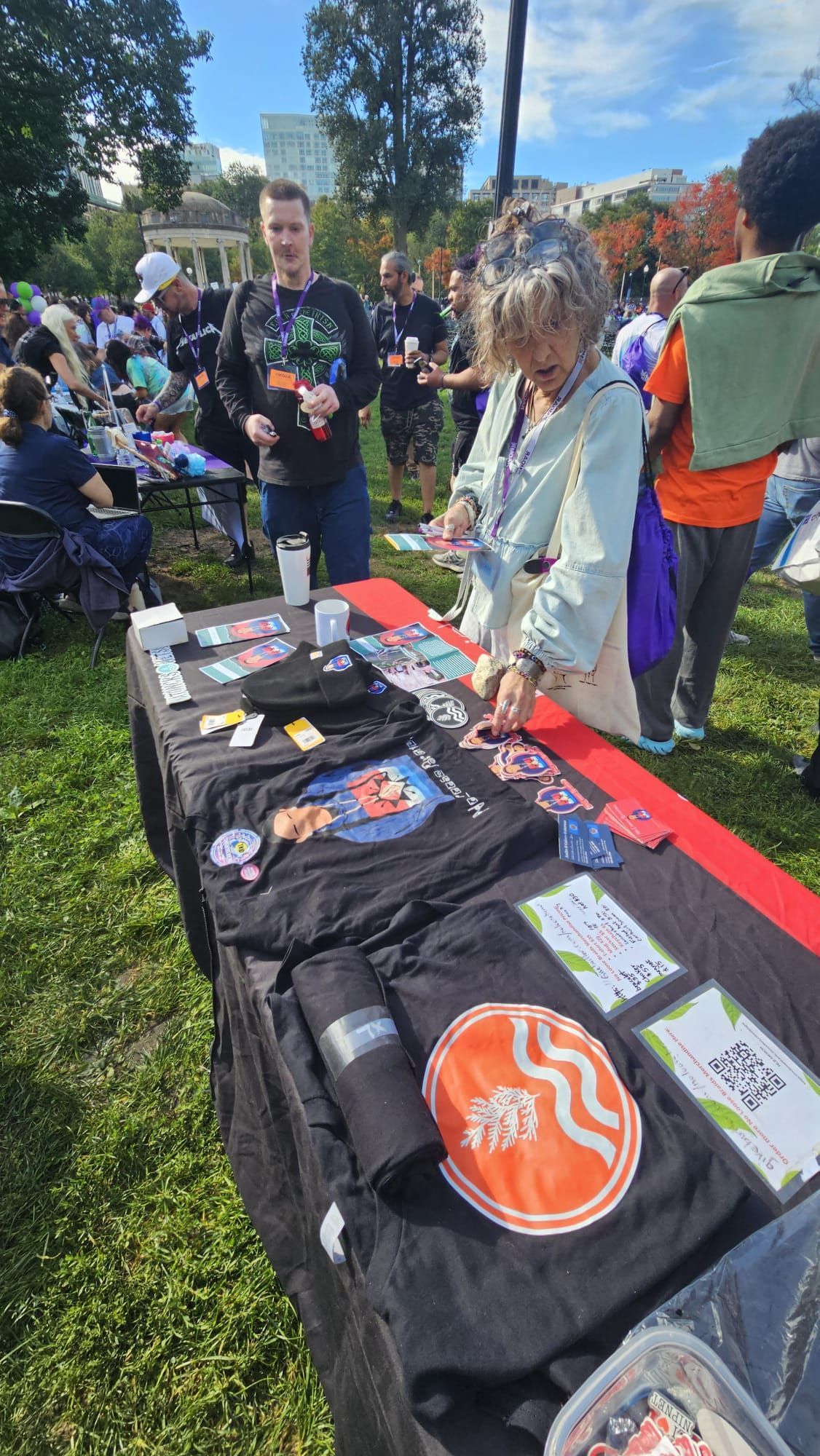A woman browses merchandise at an outdoor booth. T-shirts and stickers displayed on a table. 