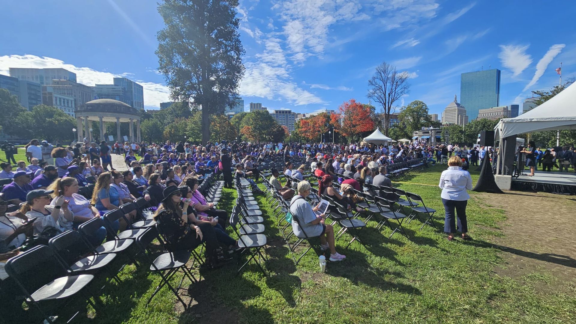 Large crowd gathered outdoors with a stage, Boston skyline in background, sunny day.