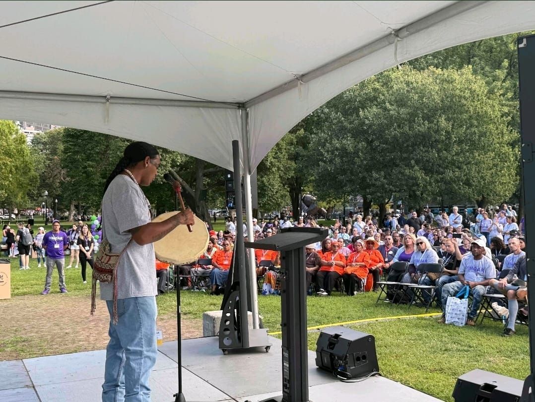 Man plays a drum on a stage outdoors. Large crowd watches, seated on chairs under a tent.
