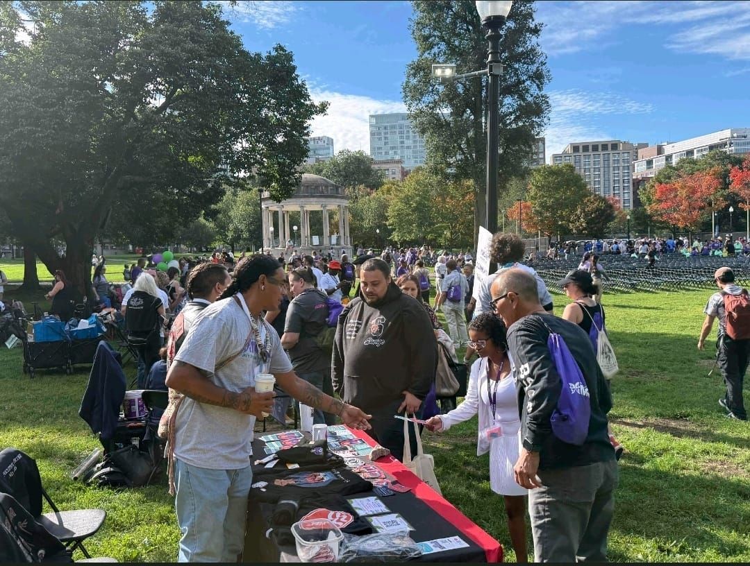People at an outdoor event with a table displaying items. Green grass, trees, and buildings are in the background.
