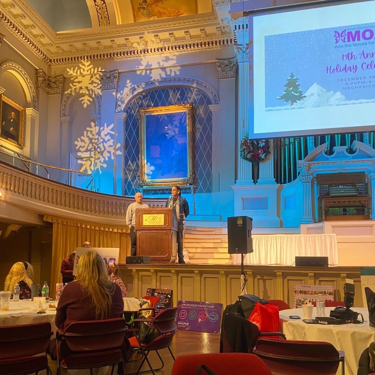 Two people on a stage giving a presentation at a holiday event in a decorated hall.