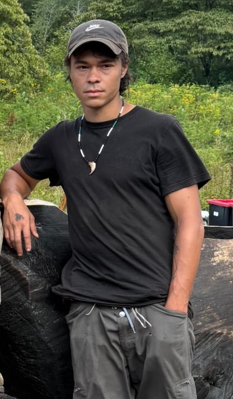 Man in black t-shirt and hat leans against a large tire. He wears a necklace and looks at the camera.