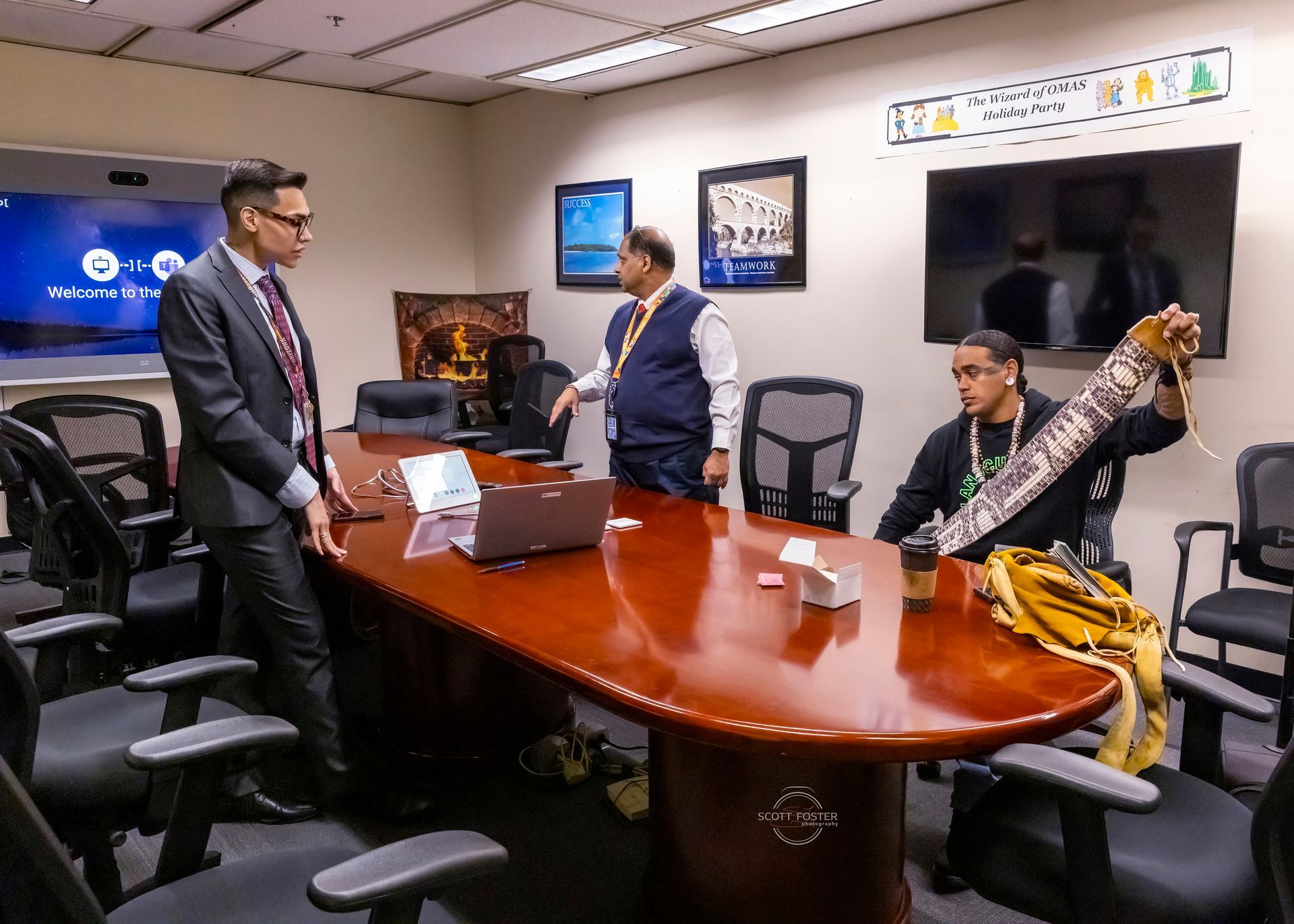 Three men in an office; one holds a beaded item, the others stand near a conference table.