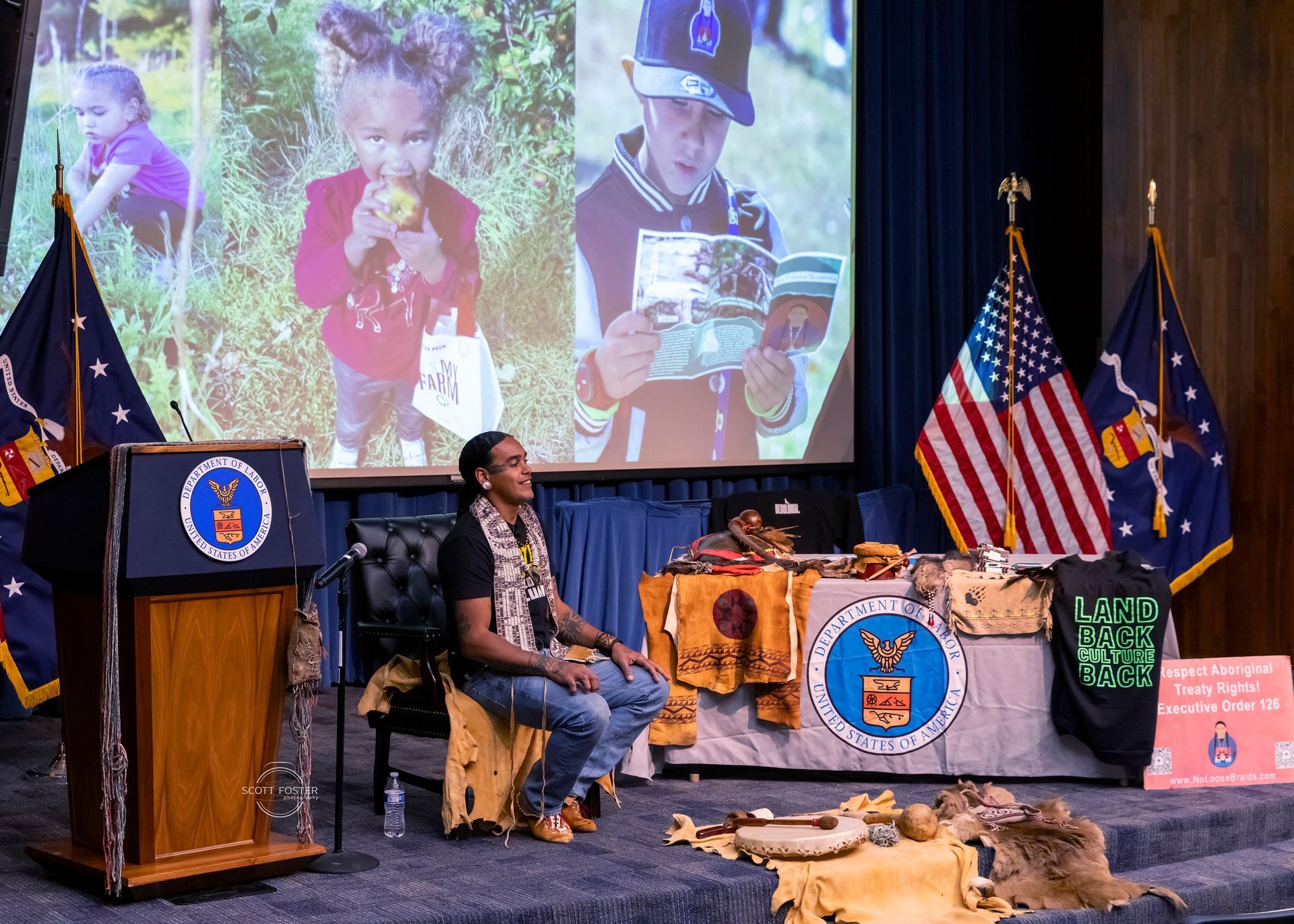 Man sits on stage near displayed artifacts with children's photos on screen, flags, podium.