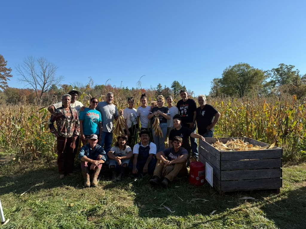 Group poses with corn harvest; sunny day in a field, wooden bin with corn, trees in the background.