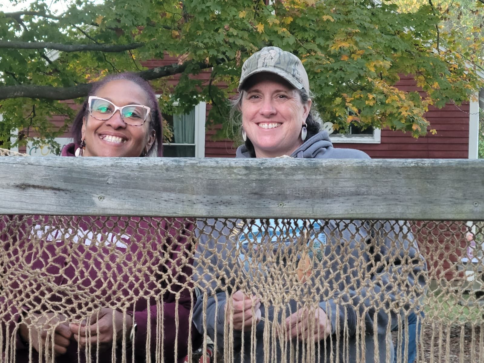 Two people smiling behind a woven rope fence, in front of a red building and trees.
