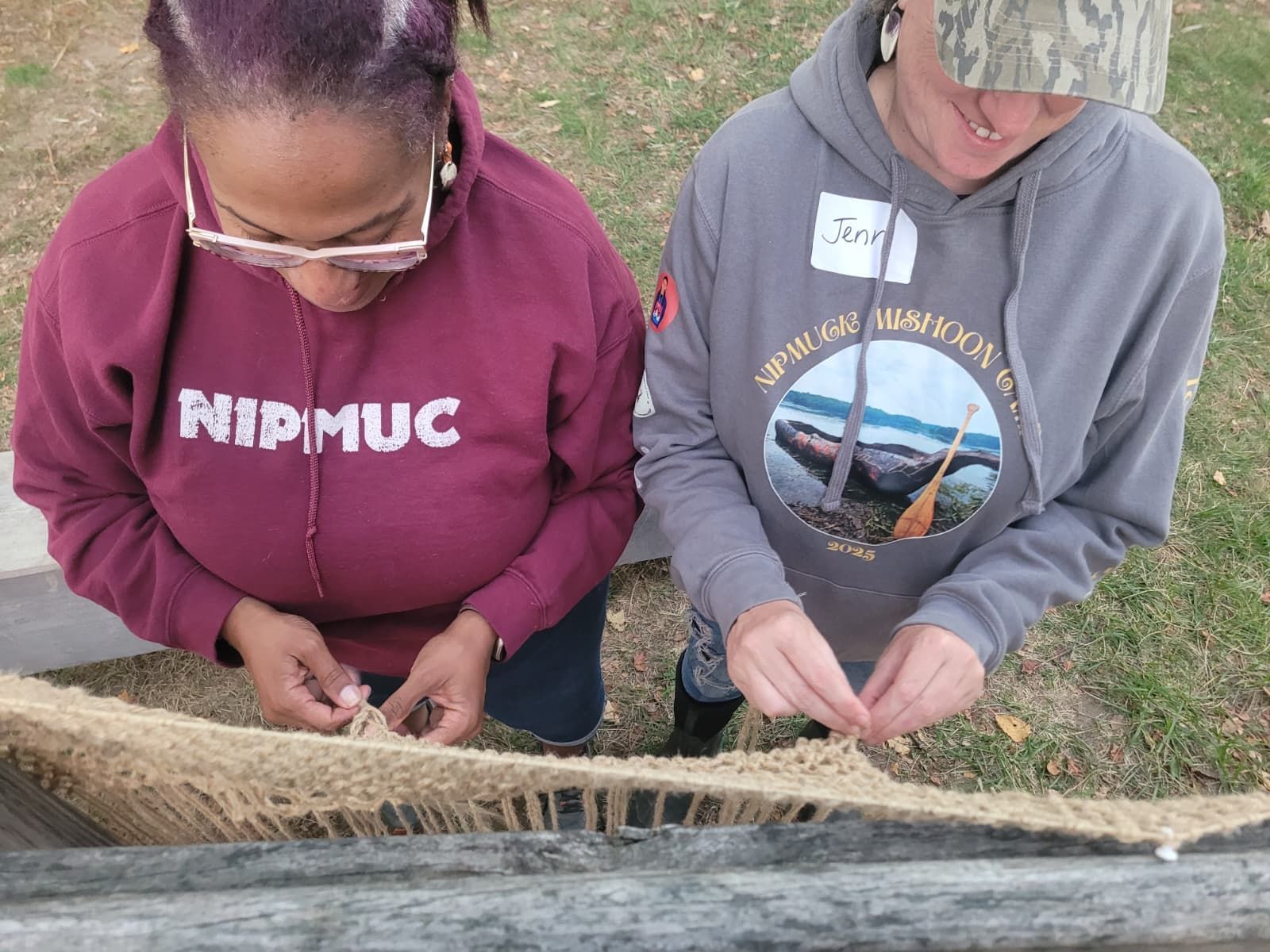 Two people weaving outdoors, focused on natural fiber material.